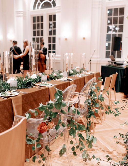 An elegant event setup with decorated tables, chairs draped with greenery, and musicians in the background in a bright, sophisticated venue.