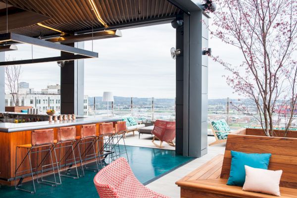 A modern rooftop bar with stylish seating, a wooden counter, and city views. Cozy atmosphere with colorful cushions and trees.