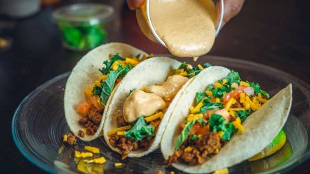 Three tacos filled with ground meat, lettuce, cheese, and tomatoes on a plate, with sauce being poured on top.