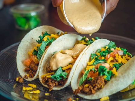 Three tacos filled with ground meat, lettuce, cheese, and tomatoes on a plate, with sauce being poured on top.