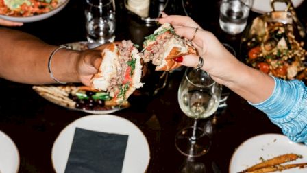 A dining table with various dishes, drinks, and people toasting with food. Fries, burgers, and seafood are visible on plates.