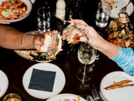 A dining table with various dishes, drinks, and people toasting with food. Fries, burgers, and seafood are visible on plates.