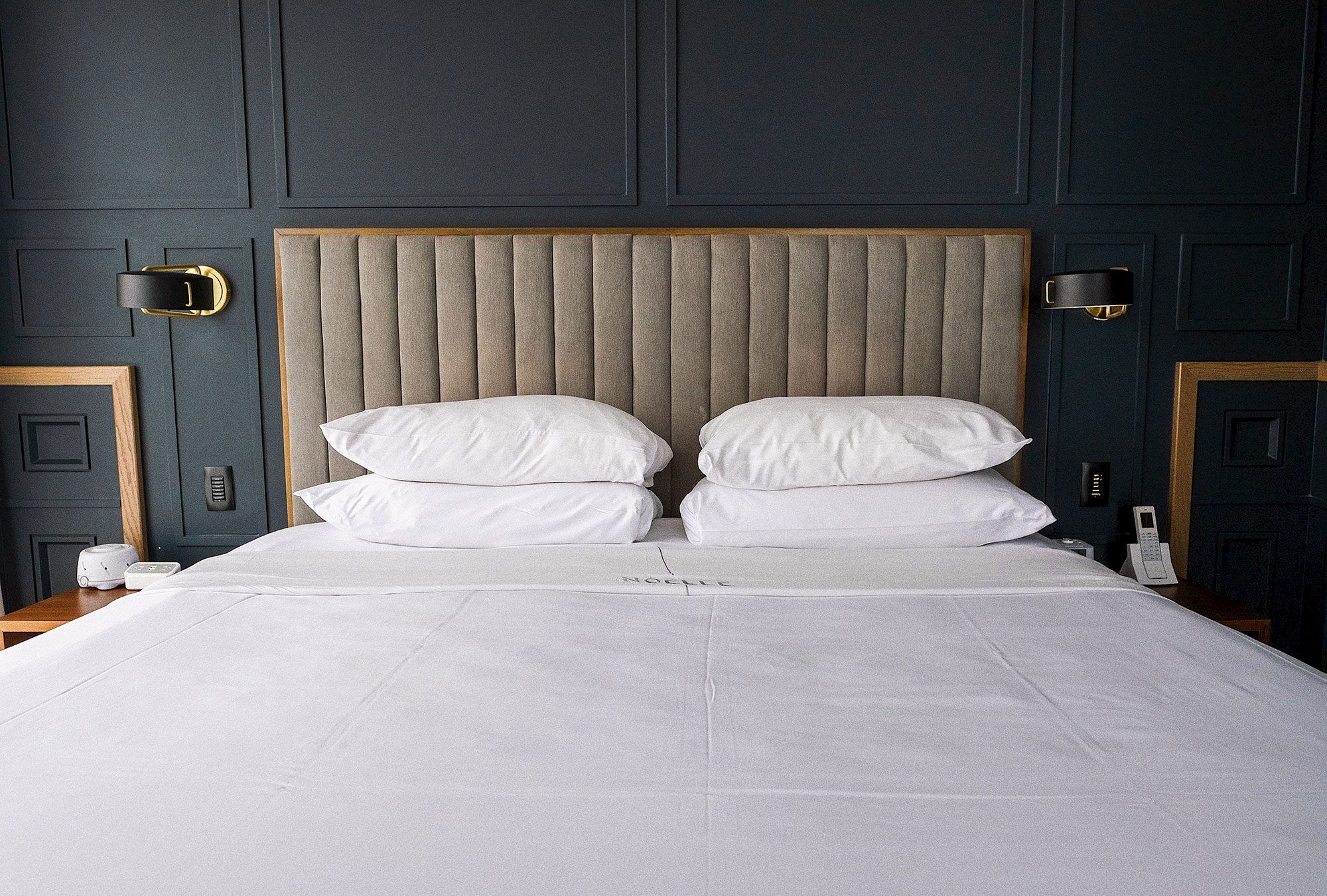 The image shows a neatly made bed with white sheets and pillows, a tufted headboard, and wall-mounted lamps on each side.