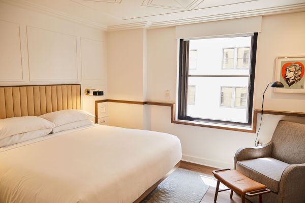 A cozy hotel room with a bed, an armchair, and a small table. There's a window and a piece of art on the wall next to the chair.