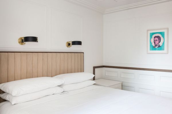 A minimalist bedroom with white walls, a wooden headboard, two pillows, and a portrait on the wall.