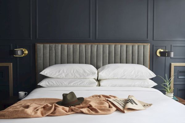 A modern bed with a grey headboard, white pillows, brown blanket, hat, and newspaper. Dark paneled wall and brass lamps complete the decor.