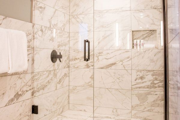 The image shows a clean, modern shower with white marble tiles and a glass door, featuring a wall-mounted showerhead and a towel hanging outside.