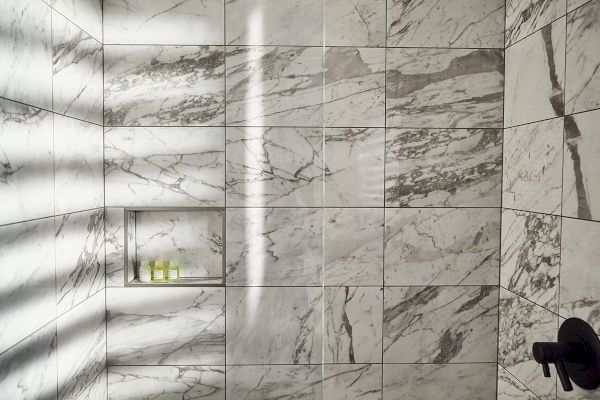 A marble-tiled shower with a niche holding small toiletries; natural light creates shadows on the walls.