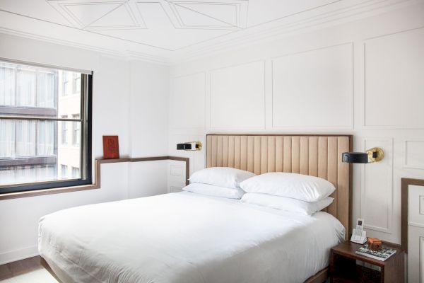 A stylish, minimalistic bedroom with a large bed, beige headboard, white bedding, wall sconces, a window, and a small bedside table.