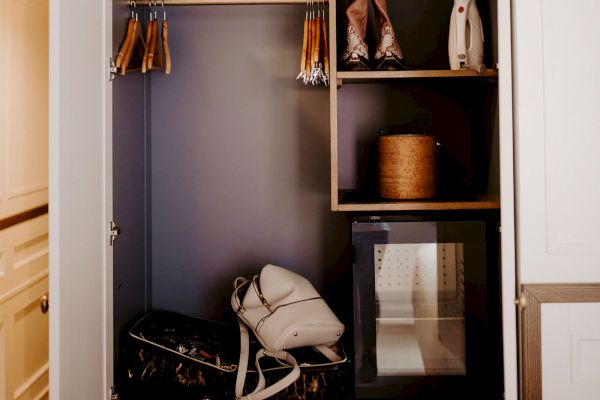 A tidy closet with hangers, shelves holding boots and a box, a backpack, and a mini-fridge or safe. Neutral-toned door slightly ajar.