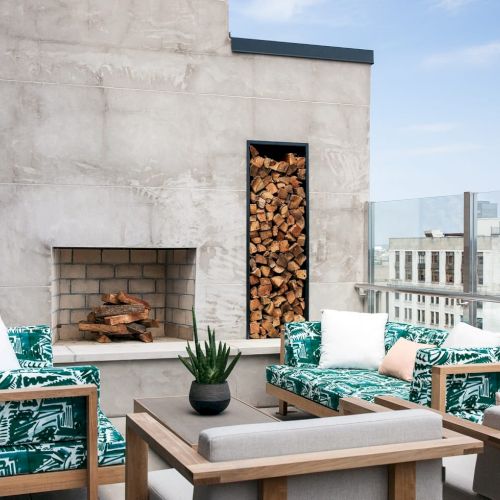 A stylish outdoor patio with green-patterned seating, a fireplace, wood stacks, and a view of city buildings under a clear sky.