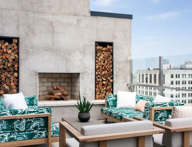 A stylish outdoor patio with green-patterned seating, a fireplace, wood stacks, and a view of city buildings under a clear sky.