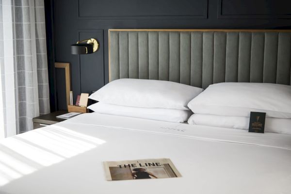 The image shows a neatly made bed with a modern headboard, wall lamp, and magazine on top, creating a cozy and stylish bedroom setting.