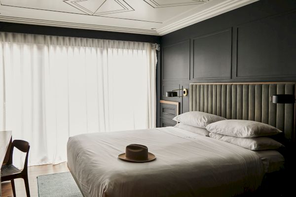 A stylish bedroom with a large bed, white linens, a hat, a chair, soft lighting, dark walls, and sheer curtains covering a window.