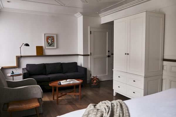 A cozy room with a dark sofa, white wardrobe, round table, chair, wall art, and a bed with a blanket.