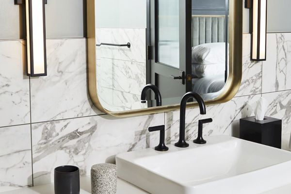 A modern bathroom features a marble wall, a large mirror, black faucets, and elegant lighting fixtures.