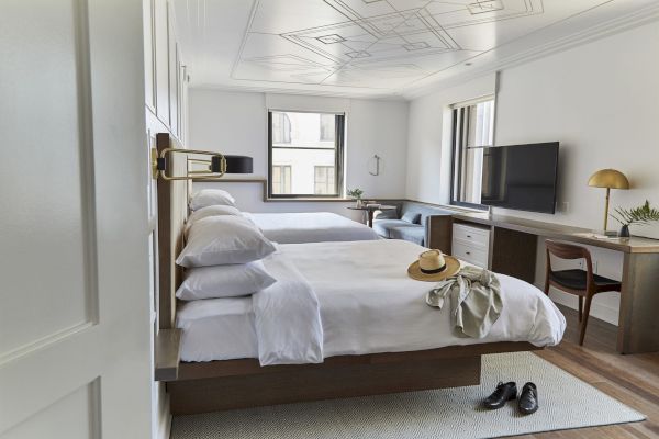 The image shows a modern hotel room with two beds, a TV, a desk, and a chair. A hat and clothing are on one bed, and shoes are on the floor.