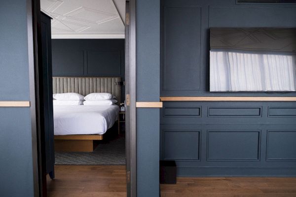 The image shows a modern bedroom with a neatly made bed, blue paneled walls, and wooden flooring, viewed through an open sliding door.