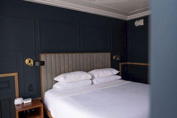 A neatly made bed with white pillows and sheets, set against a dark-colored wall with modern lamps and a side table in the corner.