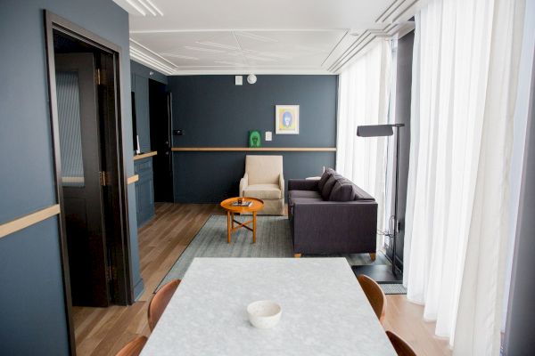 A modern living room with a sofa, table, lamp, and artwork on dark walls, paired with a dining table. White curtains filter daylight softly.