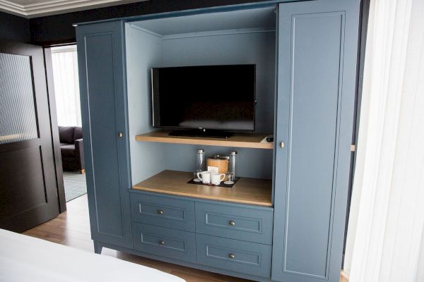 A blue entertainment cabinet with a TV, coffee setup, and shelves; it stands on a wooden floor in a room with partial bed and sofa views.