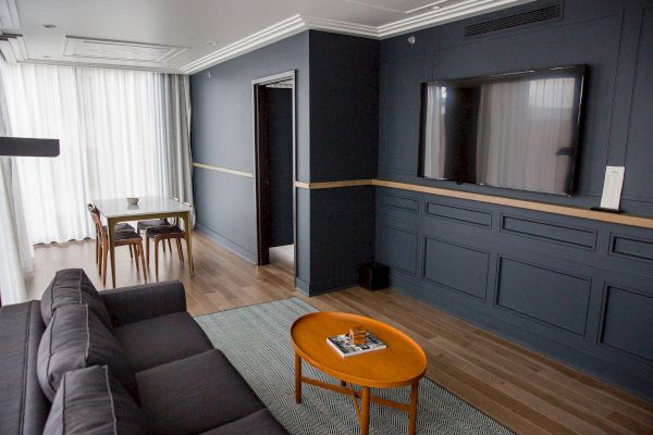 A modern living room with a gray sofa, wooden coffee table, mounted TV, dining table, and large windows covered by sheer curtains.