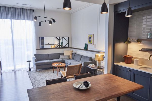 A modern living area with a gray couch, wall art, and a wooden dining table. The kitchen has dark cabinets and stylish lighting fixtures.