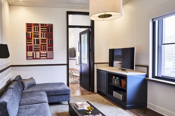 A modern living room with a gray sectional sofa, TV, abstract wall art, and large windows, featuring a stylish hanging light.