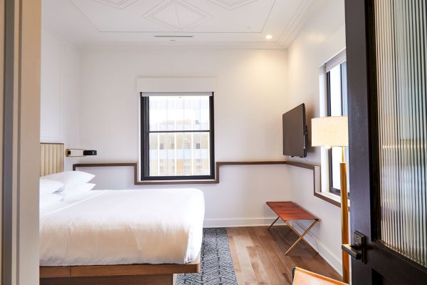 The image shows a modern, minimalistic hotel room with a bed, window, wall-mounted TV, lamp, and small bench.