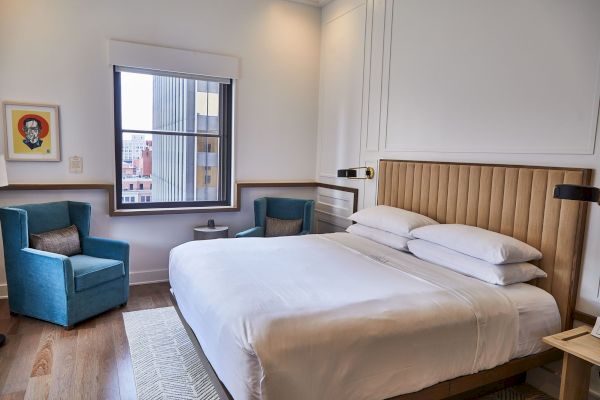 The image shows a modern hotel room with a large bed, two blue chairs, a window, and a framed picture on the wall.