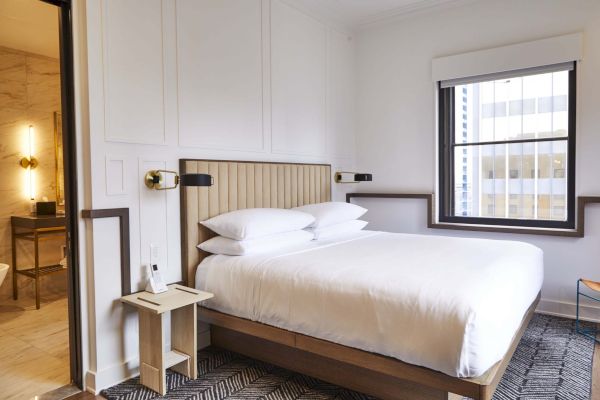 A modern hotel room with a neatly made double bed, side table, and adjoining bathroom. The window shows an urban view.