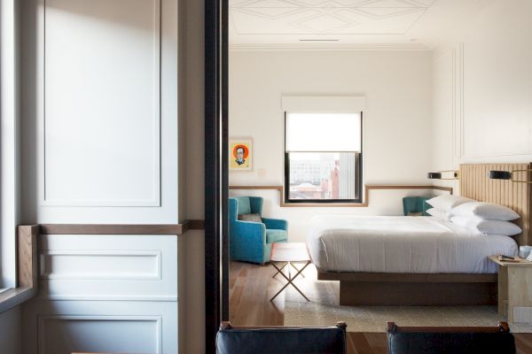 A spacious, modern bedroom with a neatly made bed, blue chair, window, and wooden accents, creating a cozy and inviting ambiance.