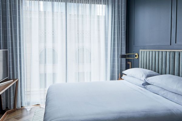 A modern bedroom with a neatly made bed, soft blue walls, a bedside light, and sheer curtains over large windows.