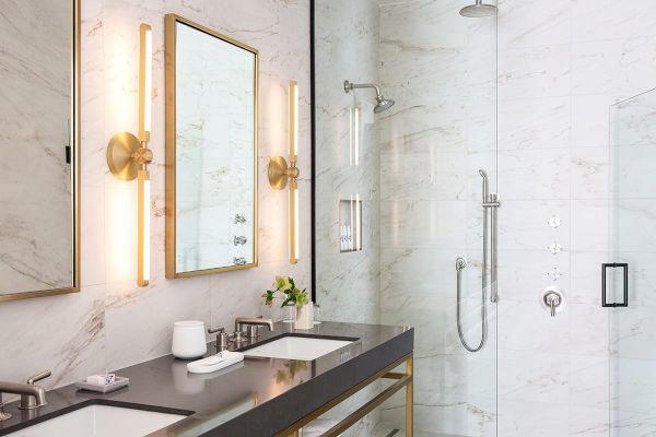 This image shows a modern bathroom with marble walls, double sinks, gold-framed mirrors, and a glass-enclosed shower area.