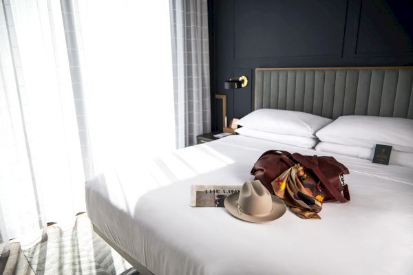 A neatly made bed with a hat, book, and scarf on it, in a well-lit room with a window and curtains.