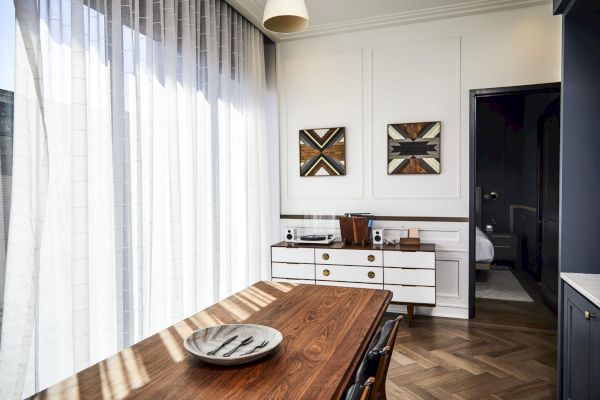 A modern dining room features a wooden table, artistic wall decor, a white cabinet, and natural light from large windows with sheer curtains.