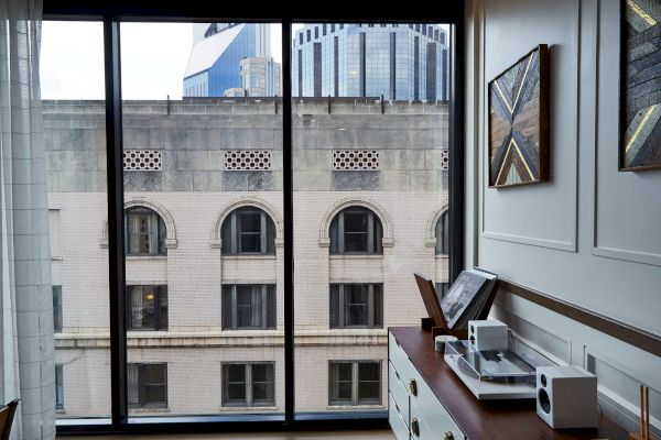 A room with a large window view of buildings; inside, a sideboard with audio equipment and framed art on the wall is visible.