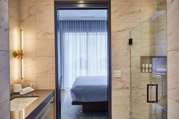 A bathroom with marble walls, a glass shower, and a view into a bedroom with curtains drawn.