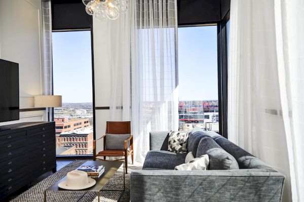 Modern living room with a gray sofa, armchair, large windows, sheer curtains, and city view. A lamp and table with decor are present.