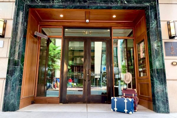 The image shows a hotel entrance with wooden doors and green marble trim, featuring suitcases and a hat placed beside the door.