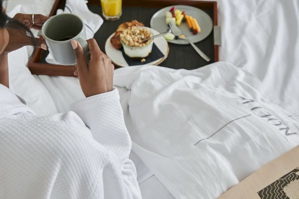 A person in a robe enjoys breakfast in bed, with coffee, juice, and fruit on a tray, reading a newspaper.