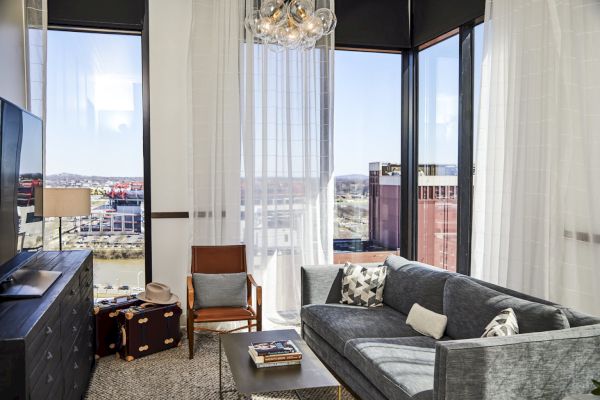 A modern living room with a gray sofa, a chair, TV, and suitcase. Large windows with sheer curtains offer a city view outside.