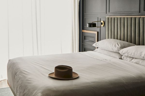 A modern bedroom with a bed, a hat on the duvet, elegant lighting, and a partially visible desk chair by the window.