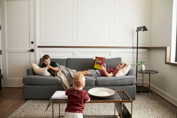 A cozy living room with three kids. One reads on a couch, another plays with a toy, and a toddler stands by a coffee table.