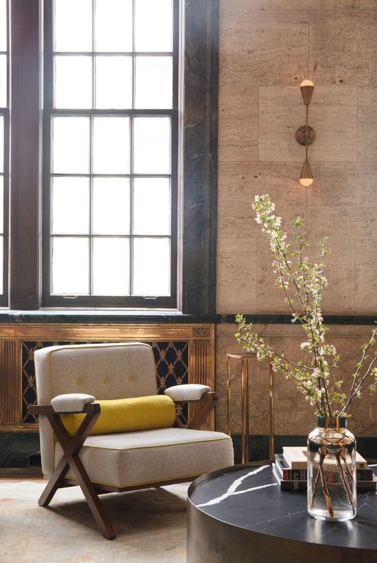 A cozy room with a beige chair, black and yellow cushion, large windows, a round table with decor, and wall lighting.