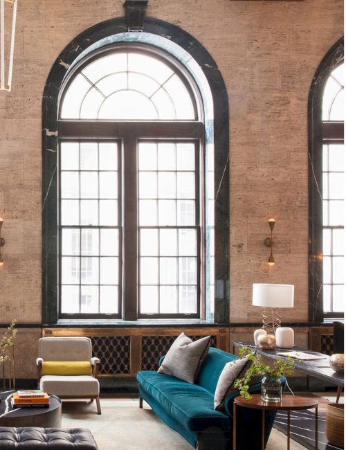 A stylish living room with large arched windows, a teal sofa, armchair, coffee table, and decor including lamps and plants.