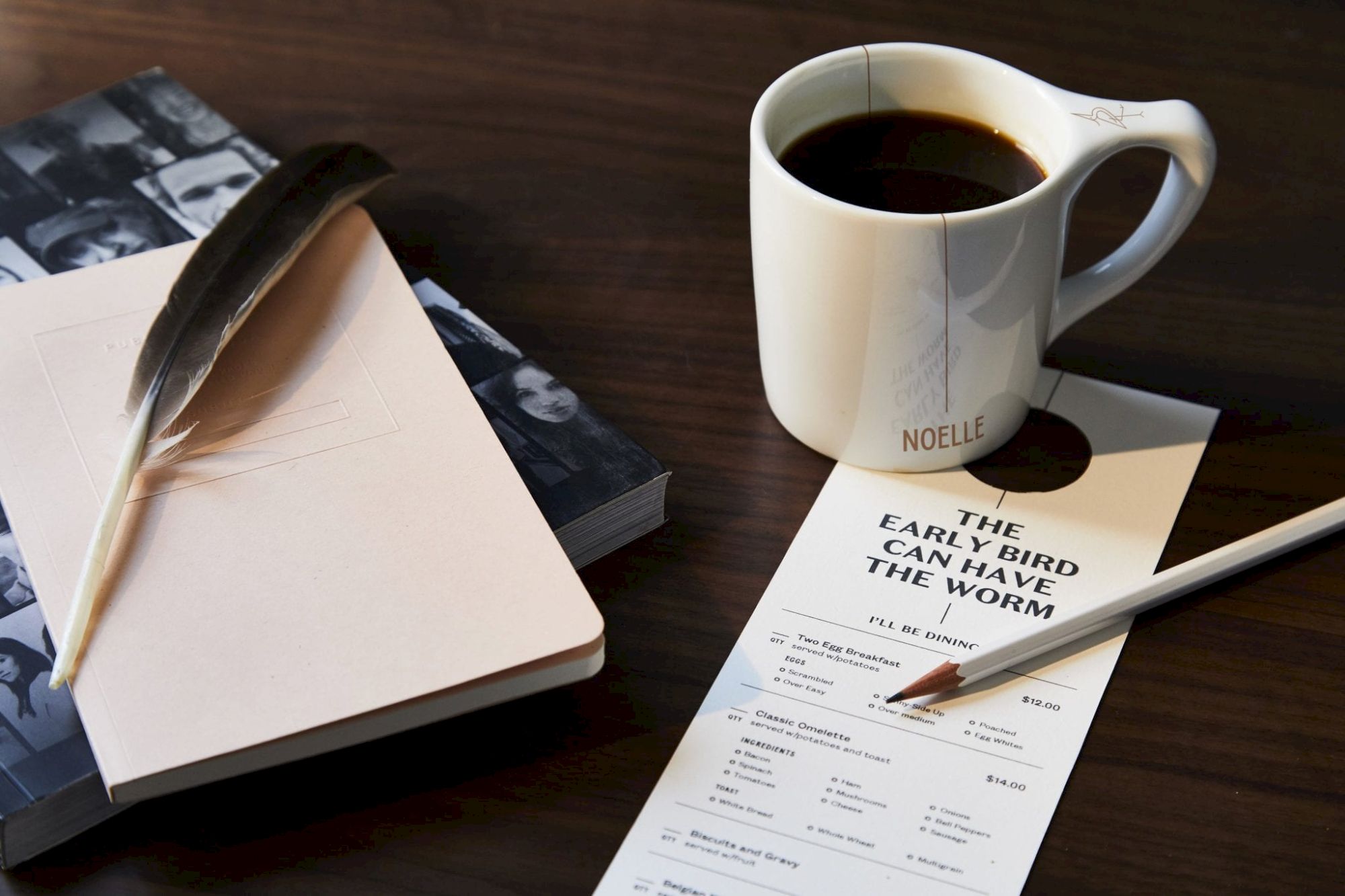 A table with a coffee cup, menu, pencil, feather, notebook, and partially visible photo book is shown.