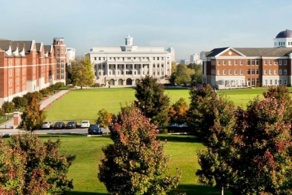 The image shows a campus with multiple brick buildings, surrounded by green lawns and trees, under a clear blue sky.