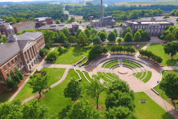 Aerial view of a university campus with green lawns, pathways, trees, and a circular amphitheater surrounded by academic buildings and nature.