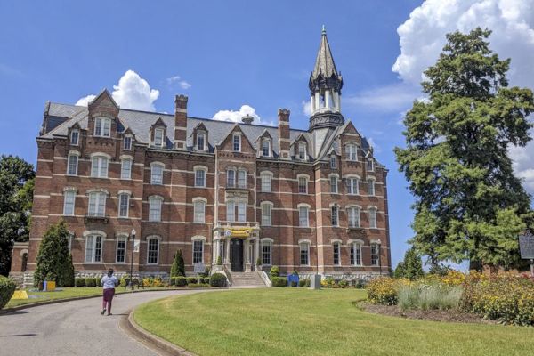 A large, historic brick building with a tall spire, surrounded by greenery and a person walking on a path in front of it.